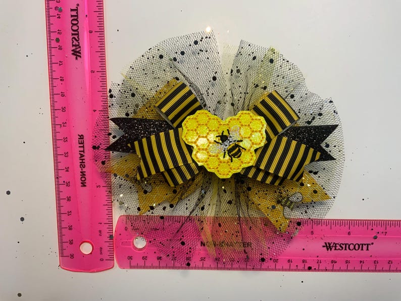 May include: A decorative hair bow featuring a bee theme. The bow has black and yellow striped ribbon, a yellow honeycomb heart with a bee, and a black tulle base with glitter accents. The bow measures approximately 4 inches in diameter.