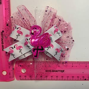 May include: A pink hair bow featuring a sequined flamingo and a white ribbon printed with pink flamingos. The bow is accented with pink glitter tulle. The bow measures approximately 4 inches.