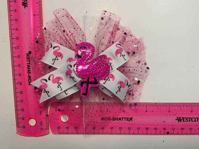 May include: A pink hair bow featuring a sequined flamingo and a white ribbon printed with pink flamingos. The bow is accented with pink glitter tulle. The bow measures approximately 4 inches.