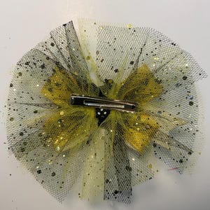 May include: A decorative hair clip with a layered design. It features yellow and black tulle, with gold glitter accents. The clip has a silver metal barrette clasp. This accessory is designed for adding flair to hairstyles.