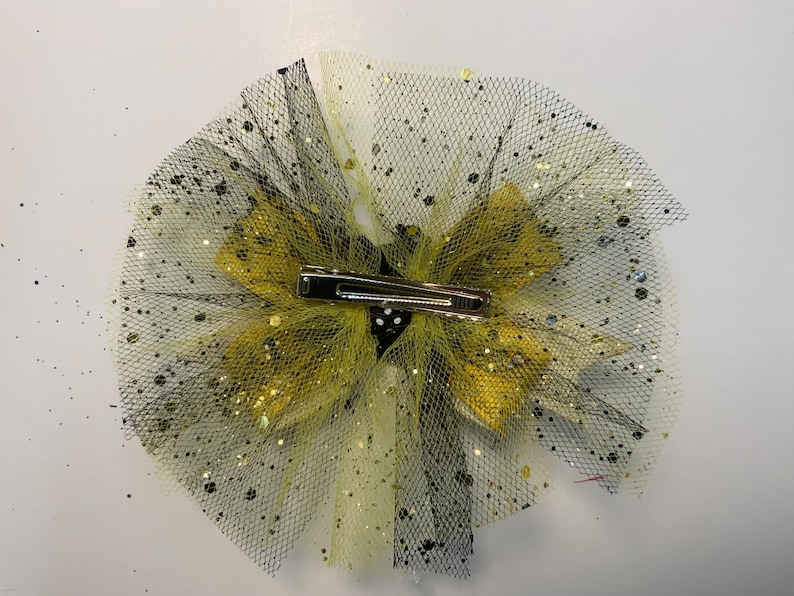 May include: A decorative hair clip with a layered design. It features yellow and black tulle, with gold glitter accents. The clip has a silver metal barrette clasp. This accessory is designed for adding flair to hairstyles.