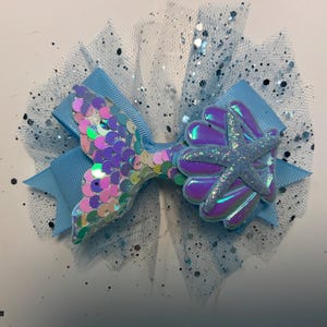 May include: A light blue hair bow with a mermaid tail and starfish design. The mermaid tail is made of iridescent sequins, and the starfish is covered in glitter. The bow is set on a layer of blue tulle with silver glitter.