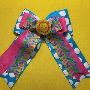 May include: A cheerful hair bow featuring a blue polka dot base with pink and patterned ribbons. The center is adorned with a yellow smiling sun embellishment. The bow is designed for a fun, summery look.