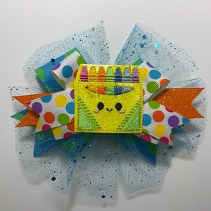 May include: A colorful hair bow featuring a yellow crayon box with a smiling face, surrounded by polka dot ribbons in various colors and orange accents. The bow is set against a light blue tulle background.