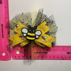 May include: A handmade hair bow featuring a yellow ribbon with bee and honeycomb patterns. The bow has a black and yellow glittery bee embellishment and is set on a black tulle base. The bow measures approximately 7.6 cm.