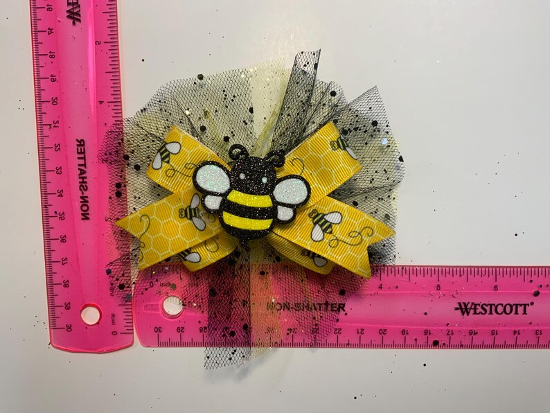 May include: A handmade hair bow featuring a yellow ribbon with bee and honeycomb patterns. The bow has a black and yellow glittery bee embellishment and is set on a black tulle base. The bow measures approximately 7.6 cm.