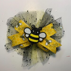 May include: A decorative hair bow with a yellow ribbon, honeycomb pattern, and bee motifs. It has a black tulle base with glitter and a bee-shaped centrepiece. A charming accessory.