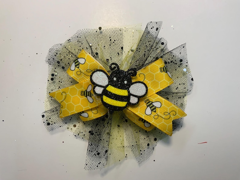 May include: A decorative hair bow with a yellow ribbon, honeycomb pattern, and bee motifs. It has a black tulle base with glitter and a bee-shaped centrepiece. A charming accessory.