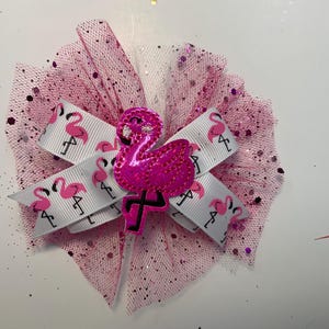 May include: A pink hair bow featuring a white ribbon with pink flamingo patterns. The bow is accented with pink tulle and a sparkly pink flamingo embellishment. The bow is designed for hair accessories.