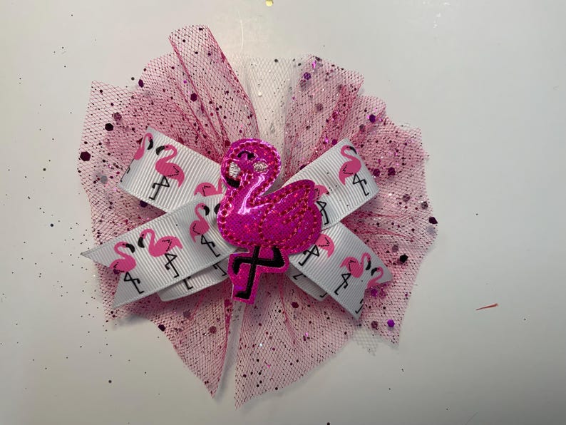 May include: A pink hair bow featuring a white ribbon with pink flamingo patterns. The bow is accented with pink tulle and a sparkly pink flamingo embellishment. The bow is designed for hair accessories.