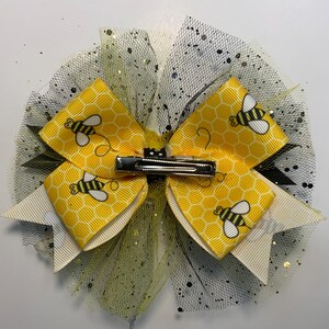 May include: A decorative hair bow featuring yellow and white ribbons with a honeycomb pattern and cartoon bees. The bow is accented with black and yellow tulle and has a silver metal clip.