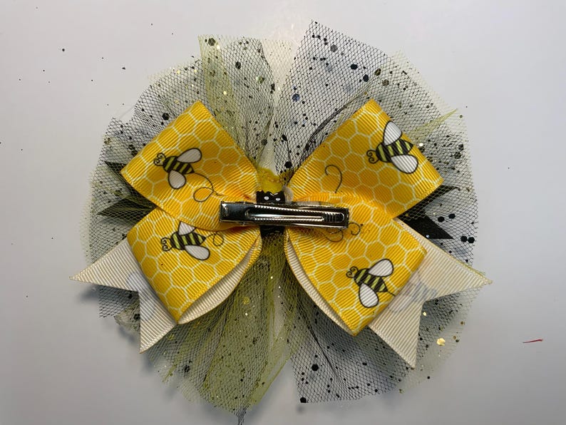 May include: A decorative hair bow featuring yellow and white ribbons with a honeycomb pattern and cartoon bees. The bow is accented with black and yellow tulle and has a silver metal clip.