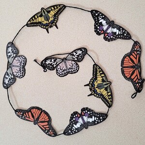 May include: A string of colourful paper butterflies, each with a unique design and colour scheme. The butterflies are strung together with black thread and are hanging against a light beige background.