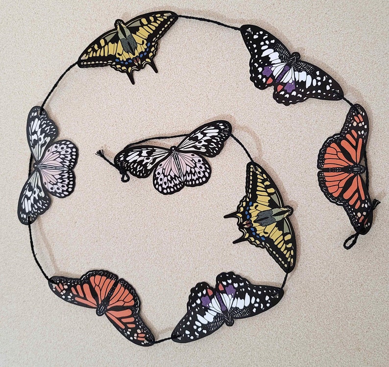 May include: A string of colourful paper butterflies, each with a unique design and colour scheme. The butterflies are strung together with black thread and are hanging against a light beige background.