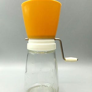 May include: A manual kitchen grinder featuring a bright yellow plastic hopper, a white plastic collar, and a clear glass jar base. A metal hand crank with a white plastic knob is attached to the side. The glass jar has embossed measurement markings.