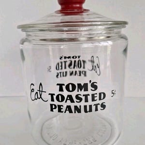 May include: A clear glass jar with a red lid featuring the embossed word Tom's. The front of the jar displays black text reading Eat Tom's Toasted Peanuts 5 cents. The glass is transparent and cylindrical with a wide opening and a matching glass lid.