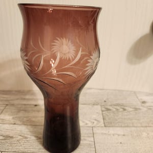 May include: A tall, elegant, purple glass vase with a flared top and a slender base. The vase features etched floral designs. The glass transitions from a lighter purple at the top to a darker shade at the base. A decorative piece for the home.
