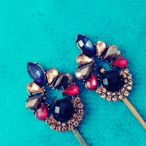 Puede incluir: Dos horquillas decorativas con adornos de joyas. Cada horquilla presenta un diseño circular con gemas azules, rosas y negras sobre una base dorada. El fondo es de color turquesa sólido. Accesorios para el cabello.