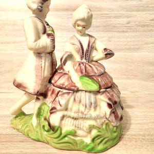 May include: A ceramic figurine depicting a couple in period attire. The figure on the left wears a light-colored coat with purple accents, while the figure on the right wears a layered purple dress with a green leaf-shaped detail, all on a green base.