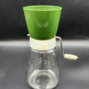 May include: A manual kitchen grinder featuring a bright green plastic hopper, a white plastic collar, and a clear glass base container. A metal hand crank with a small white handle is attached to the side for operation of the grinding mechanism.