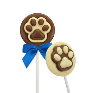 May include: Two chocolate lollipops featuring paw print designs. One is dark chocolate with a light brown paw print and a blue ribbon. The other is white chocolate with a dark brown paw print. Both are on white sticks.