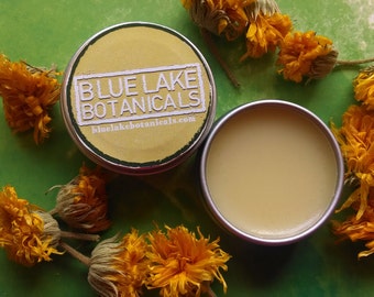May include: A small tin container with a yellow lid and label that reads "BLUE LAKE BOTANICALS" and "bluelakebotanicals.com". The tin is open and a small round container of yellow balm is visible. The background is a green surface with yellow flowers.