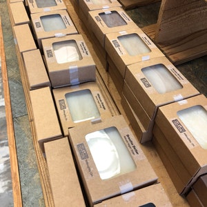 May include: A display of natural vegan soap bars in brown cardboard boxes with clear windows showing the soap inside. The soap bars are labeled with different scents, including Sweet Almond, Lavender, Charcoal & Clay, and Breathe Better. The boxes are stacked on wooden shelves.