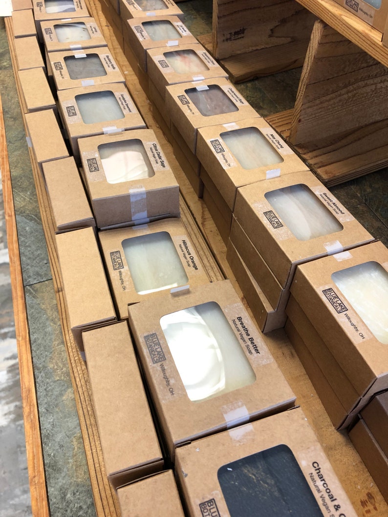 May include: A display of natural vegan soap bars in brown cardboard boxes with clear windows showing the soap inside. The soap bars are labeled with different scents, including Sweet Almond, Lavender, Charcoal & Clay, and Breathe Better. The boxes are stacked on wooden shelves.