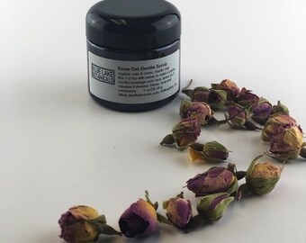 May include: A black jar of Rose Oat Gentle Scrub by Blue Lake Botanicals. The jar is sitting on a white surface with dried rose buds scattered around it. The label on the jar says "Rose Oat Gentle Scrub" and lists the ingredients as organic oats & roses.  It also says "Apply a pea-sized amount to wet skin. Gently massage with wet fingertips. Rinse thoroughly.  Use 1-2 times per week.  Store in a cool, dry place. 1 fl oz / 30 ml.  BlueLakeBotanicals.com.  Wilmington, DE."