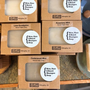 May include: Five brown cardboard boxes with clear windows showing white bars of soap. Each box has a label with the name of the soap scent, "Skin, Hair & Beard Shampoo Soap", and the brand name "Blue Lake Botanicals". The scents are Insect Repellent, Rosemary Lavender, Lime Eucalyptus, Rosemary Mint, and Cedarwood Mint.