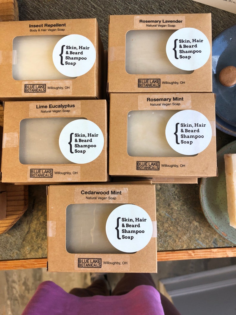 May include: Five brown cardboard boxes with clear windows showing white bars of soap. Each box has a label with the name of the soap scent, "Skin, Hair & Beard Shampoo Soap", and the brand name "Blue Lake Botanicals". The scents are Insect Repellent, Rosemary Lavender, Lime Eucalyptus, Rosemary Mint, and Cedarwood Mint.