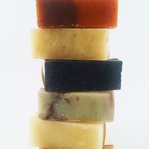 May include: A stack of six handmade soap bars in various colors and patterns. The bars are stacked on top of each other, with the top bar being a brown and orange color. The bars are all rectangular in shape and have a smooth surface.