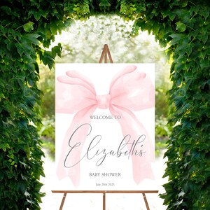May include: A white welcome sign with a large pink watercolor bow. The text reads "Welcome to Elizabeth's Baby Shower July 28th 2025".
