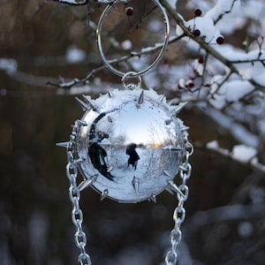 May include: A reflective silver sphere ornament with sharp spikes, suspended by a chain and ring. The sphere reflects a snowy outdoor scene. The ornament hangs from a snow-covered branch, creating a unique holiday decoration.
