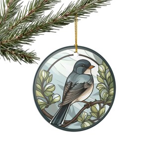 Può includere: Ornamento natalizio rotondo in ceramica con un motivo di uccello stilizzato. L'uccello ha piume blu-grigie e petto beige, posato su un ramo con foglie verdi. L'ornamento ha un bordo grigio scuro e un cordino dorato. Sfondo azzurro.