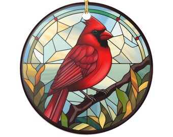 May include: A round ceramic ornament featuring a vibrant red cardinal perched on a brown branch. The bird is set against a stained-glass style background with blue, yellow, and green hues. A gold string is attached for hanging.