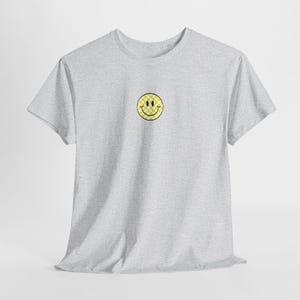 May include: A light gray t-shirt featuring a yellow and white checkered smiley face design on the chest. The smiley face has a black outline and is centered on the shirt. The shirt has short sleeves and a crew neck.