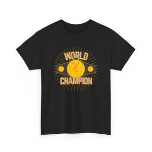 May include: Black t-shirt featuring a gold and yellow graphic of a championship belt. The belt has the words "WORLD CHAMPION" in gold lettering. The design includes a central emblem and two side emblems.