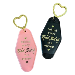 May include: Two keychains shaped like motel room keys. One is pink with gold lettering that says "behind every Bad Bitch is a carseat". The other is black with gold lettering that says "behind every Bad Bitch is a carseat".