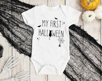 Puede incluir: Body de bebé blanco con el texto negro "MY FIRST HALLOWEEN". El diseño incluye un gato negro, una telaraña, murciélagos y estrellas. En el fondo hay calabazas decorativas, arañas y tela negra.