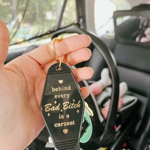 May include: Black keychain shaped like a motel key with gold lettering that reads "behind every Bad. Bitch is a carseat" with a heart symbol before and after the text.