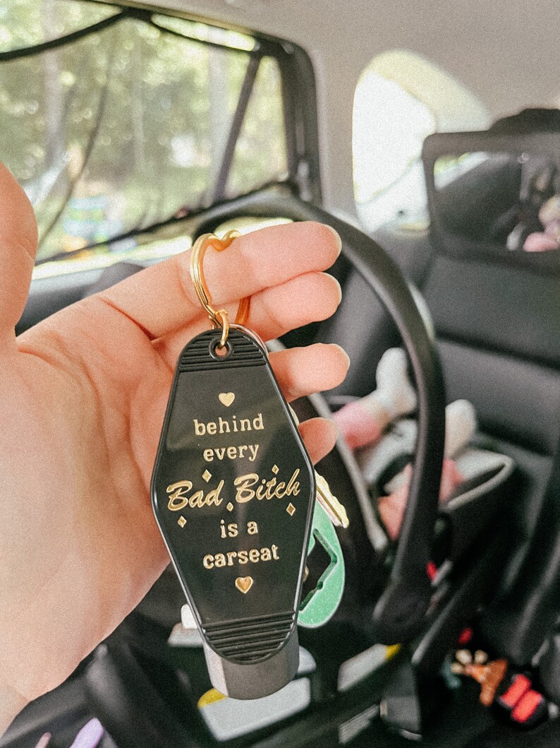 May include: Black keychain shaped like a motel key with gold lettering that reads "behind every Bad. Bitch is a carseat" with a heart symbol before and after the text.