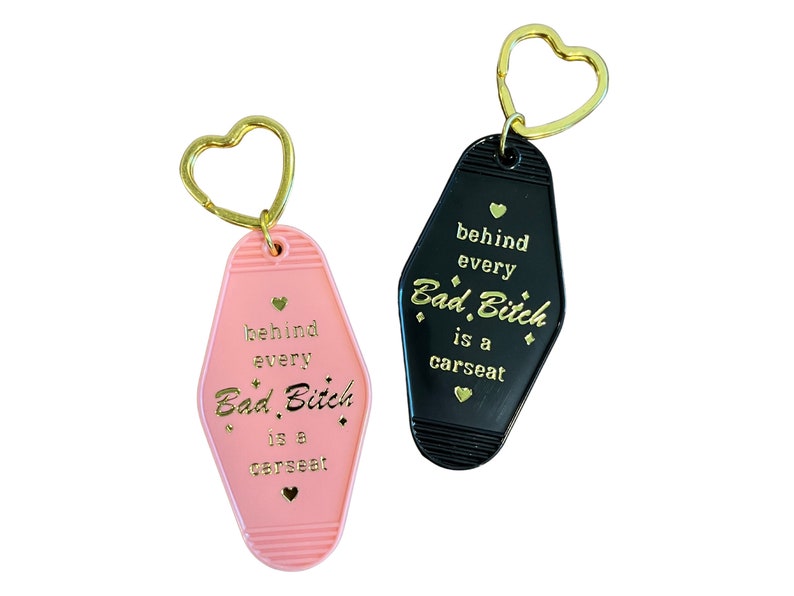 May include: Two keychains shaped like motel room keys. One is pink with gold lettering that says "behind every Bad Bitch is a carseat". The other is black with gold lettering that says "behind every Bad Bitch is a carseat".