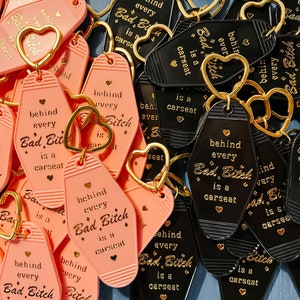 May include: A collection of pink and black keychains with the text "Behind every Bad Bitch is a carseat" in gold lettering. The keychains are in the shape of motel keys.