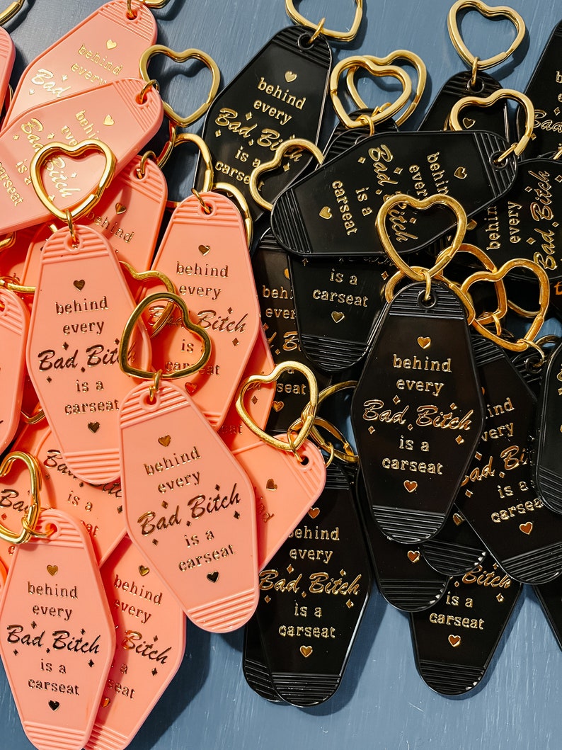 May include: A collection of pink and black keychains with the text "Behind every Bad Bitch is a carseat" in gold lettering. The keychains are in the shape of motel keys.