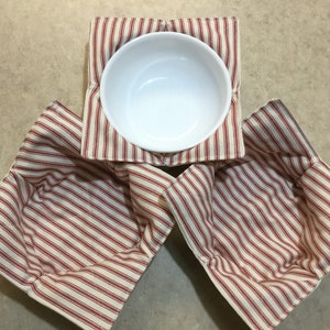 May include: A white ceramic bowl rests on a red and white striped fabric bowl cozy. Two additional bowl cozies are visible, also with red and white stripes. The cozies are designed to hold bowls and protect surfaces.
