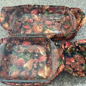 May include: Set of three glass baking dishes with colorful fabric covers. The fabric features a pattern of pumpkins, apples, and leaves in shades of orange, red, and green on a dark blue background. The set includes two rectangular dishes and one square dish.