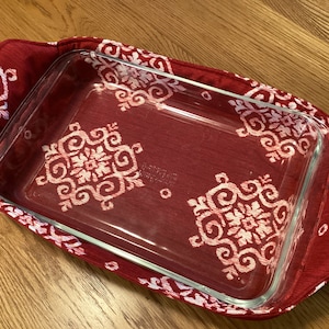 May include: A rectangular glass baking dish rests inside a red fabric carrier. The carrier features a white and red snowflake pattern. The dish is on a wooden surface.