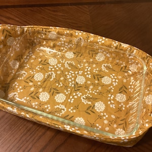 May include: A yellow fabric casserole dish carrier with a white floral pattern. The carrier has handles on the sides and is holding a clear glass baking dish.
