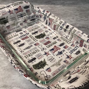 May include: A rectangular glass baking dish nestled inside a fabric hot pad. The fabric features a patriotic print with American flags, eagles, and text including Courage, Loyalty, Strength, Respect, Valor, and Land of the Free on a white background.